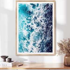 May include: A rectangular wall art print featuring an aerial view of ocean waves crashing, displaying a gradient of deep navy blue to bright turquoise water with white sea foam, enclosed in a light-toned wooden frame with a white mat border.