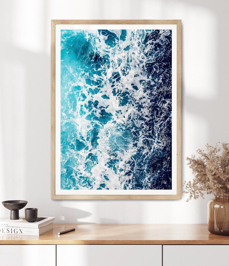 May include: A rectangular wall art print featuring an aerial view of ocean waves crashing, displaying a gradient of deep navy blue to bright turquoise water with white sea foam, enclosed in a light-toned wooden frame with a white mat border.
