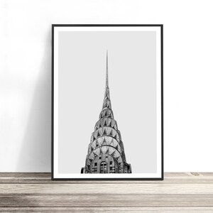 May include: A black and white framed art print featuring the iconic tiered, sunburst-patterned spire of the Chrysler Building in New York City. The architectural photograph captures the skyscraper's distinctive metallic crown against a light gray sky.