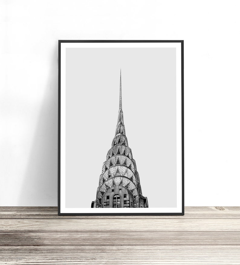 May include: A black and white framed art print featuring the iconic tiered, sunburst-patterned spire of the Chrysler Building in New York City. The architectural photograph captures the skyscraper's distinctive metallic crown against a light gray sky.