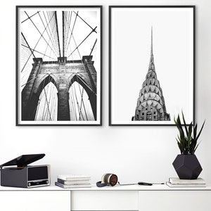 May include: Two framed black and white wall art prints featuring architectural photography. The left print displays the Brooklyn Bridge with its suspension cables and stone arches. The right print shows the spire of the Chrysler Building.