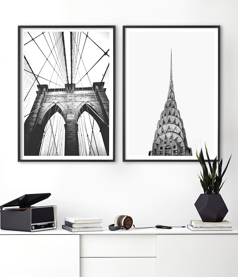 May include: Two framed black and white wall art prints featuring architectural photography. The left print displays the Brooklyn Bridge with its suspension cables and stone arches. The right print shows the spire of the Chrysler Building.