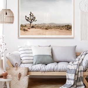 May include: A framed landscape print featuring a desert scene with a Joshua tree. The print has a light wood frame and is displayed above a sofa with pillows and a woven tote bag. The color palette is neutral with beige, brown, and gray tones.
