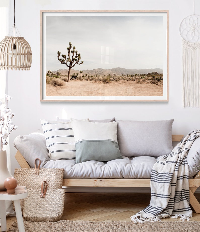May include: A framed landscape print featuring a desert scene with a Joshua tree. The print has a light wood frame and is displayed above a sofa with pillows and a woven tote bag. The color palette is neutral with beige, brown, and gray tones.