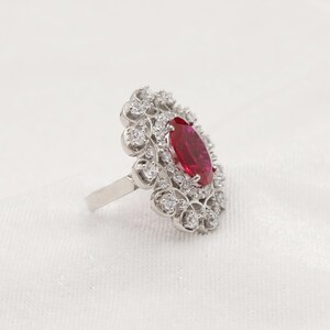 Puede incluir: Un anillo de plata con una gran piedra ovalada roja rodeada de piedras blancas m&aacute;s peque&ntilde;as. El anillo tiene un delicado dise&ntilde;o floral.