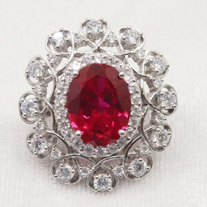 Puede incluir: Un anillo de plata con una gran piedra de rub&iacute; de forma ovalada rodeada por un halo de piedras m&aacute;s peque&ntilde;as y transparentes. El anillo presenta un delicado dise&ntilde;o floral.