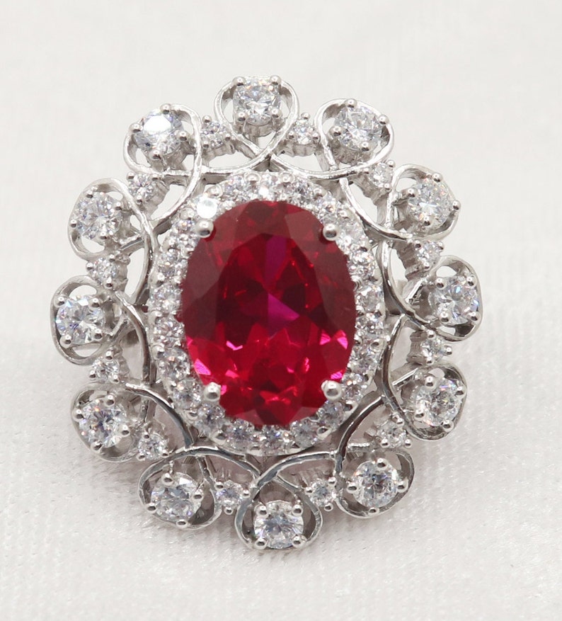 Puede incluir: Un anillo de plata con una gran piedra de rub&iacute; de forma ovalada rodeada por un halo de piedras m&aacute;s peque&ntilde;as y transparentes. El anillo presenta un delicado dise&ntilde;o floral.