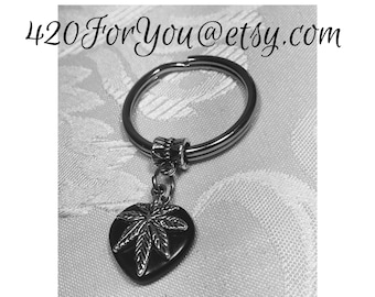 May include: A silver key chain with a black heart charm featuring a silver marijuana leaf design. The charm is attached to the key chain with a silver bead.