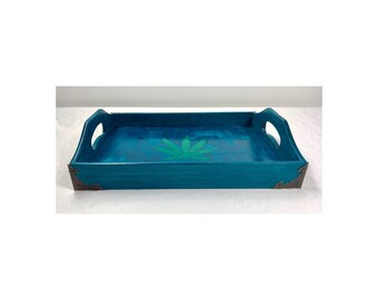 May include: A teal blue wooden serving tray with a green marijuana leaf design. The tray has two handles and silver metal corner accents.