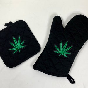 Puede incluir: Un juego de dos manoplas de horno negras con un diseño de hoja de marihuana verde. Las manoplas están acolchadas y tienen un lazo para colgar.
