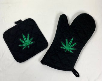 May include: A set of two black oven mitts with a green marijuana leaf design. The mitts are quilted and have a hanging loop.