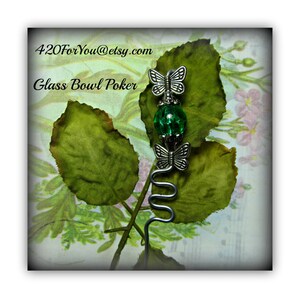 May include: A silver metal poker with a green glass bead and two silver butterfly charms. The poker is on a green leaf background with the text "420ForYou@etsy.com" and "Glass Bowl Poker" in black.