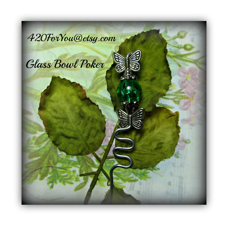 May include: A silver metal poker with a green glass bead and two silver butterfly charms. The poker is on a green leaf background with the text "420ForYou@etsy.com" and "Glass Bowl Poker" in black.