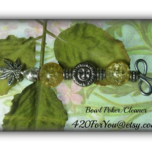 May include: A silver bowl poker/cleaner with a leaf design and a sun face charm. The poker is decorated with clear yellow glass beads. The poker is on a green and white background.