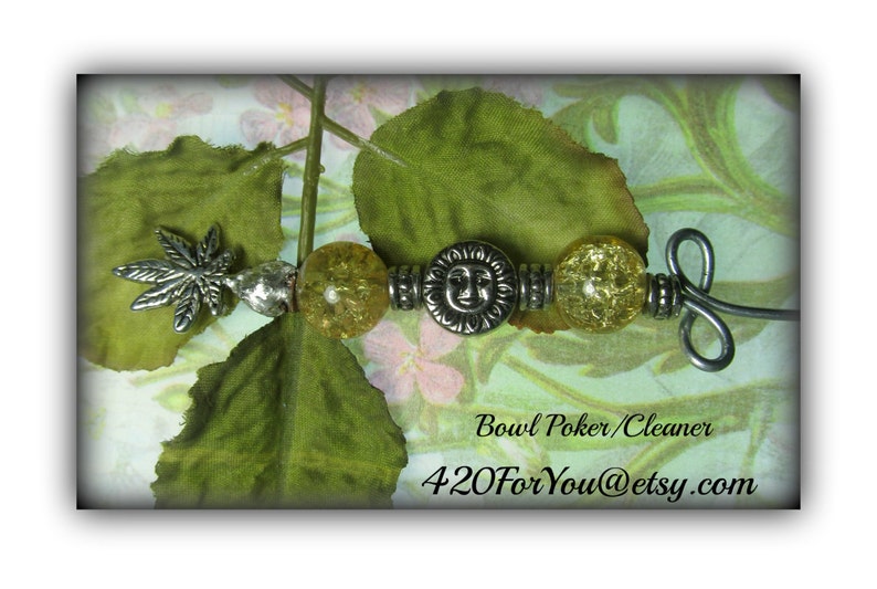 May include: A silver bowl poker/cleaner with a leaf design and a sun face charm. The poker is decorated with clear yellow glass beads. The poker is on a green and white background.