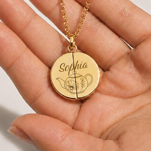 Puede incluir: Un collar dorado con un colgante redondo. El colgante está dividido en dos mitades y presenta un diseño de tetera y el nombre "Sophia" grabado. El collar está sostenido en la mano de una persona.