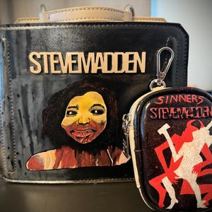May include: Black handbag and coin purse set. The handbag features the text "STEVEMADDEN" in gold lettering, with a painted portrait. The coin purse has the text "SINNERS STEVEMADDEN" and a red and white graphic.