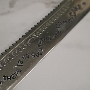Könnte beinhalten: Ein silberfarbenes Tortenmesser aus Metall mit gezackter Klinge. Die Klinge ist mit dem Text "THERE IS NO SUCH THING AS TOO MUCH CAKE" in einer geschwungenen, gestempelten Schrift graviert, die am Anfang und Ende mit kleinen Herzsymbolen verziert ist.