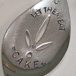 Può includere: Un cucchiaio scanalato in acciaio inossidabile con la frase "LET THEM EAT CAKE" stampata sulla superficie. Il cucchiaio presenta ritagli decorativi e simboli a forma di cuore. Un utensile da cucina unico.