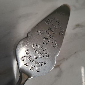 May include: A silver cake server with a curved, petal-shaped blade. The text "BIRTHDAYS ARE JUST NATURE'S WAY OF TELLING YOU TO EAT MORE CAKE" is engraved on the blade. The server has a long, slender handle.