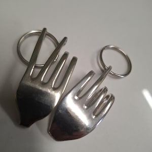 May include: Two silver fork-shaped keyrings. Each keyring has a fork head attached to a small, round ring. The forks have four tines and a polished finish, reflecting the light. The keyrings are displayed on a white surface.