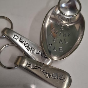 Puede incluir: Un conjunto de llaveros de metal plateado. Uno tiene forma de cuchara con el texto "REAL MEN DRINK REAL ALE". Otro dice "LAGER LOUT" con una estrella. El tercer llavero dice "BEER MONSTER".