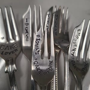 Puede incluir: Una colección de tenedores de plata con varios mensajes grabados, incluyendo "Cake Lover", "I Love Cake", "Fiona's Cake", "York Cake", "My Cake" y "Emergency Cake".