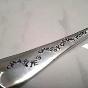 May include: A close-up shot of a silver fork with the words "CHARLIE'S CHEESY CHIPS FORK" engraved on the handle. The fork is made of shiny metal and is set against a white background. The text is in a playful, slightly uneven font.