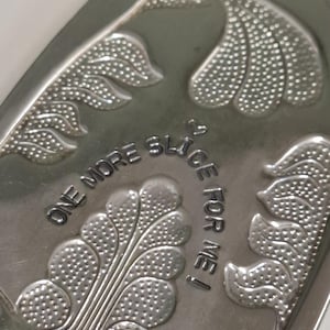 Może przedstawiać: Srebrny półmisek do serwowania z wytłoczonymi wzorami kwiatowymi i napisem "ONE MORE SLICE FOR ME!". Półmisek ma szczegółowe wzory liści i płatków, tworząc dekoracyjny i elegancki wygląd.