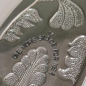 Puede incluir: Una fuente plateada con diseños de hojas en relieve y el texto "ONE MORE SLICE FOR ME!". La fuente tiene una forma ligeramente curvada y un acabado pulido, adecuada para servir pasteles u otros postres.