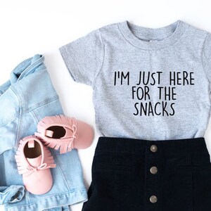 May include: A grey short-sleeved t-shirt with the text "I'm just here for the snacks" in black. The shirt is paired with a black skirt and pink shoes.