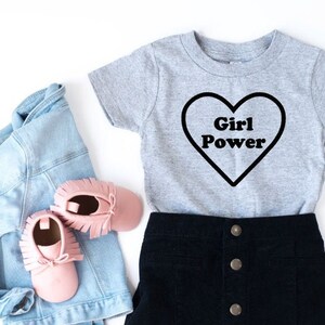 Pode incluir: Uma camiseta cinza de manga curta com um gr&aacute;fico de cora&ccedil;&atilde;o preto que diz "Girl Power". A camiseta &eacute; combinada com uma saia de veludo cotel&ecirc; preta e uma jaqueta jeans azul claro. Tamb&eacute;m est&atilde;o inclu&iacute;dos mocassins rosa na imagem.