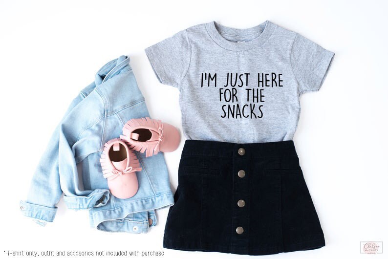 May include: A grey short-sleeved t-shirt with the text "I'm just here for the snacks" in black. The shirt is paired with a black skirt and pink shoes.