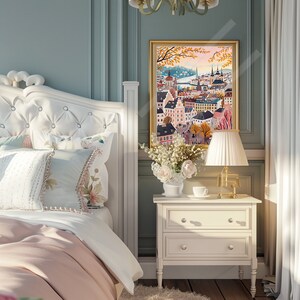 May include: A white tufted headboard with a pink and white bedding set. A white bedside table with two drawers sits next to the bed. A framed print of a city skyline with a gold frame hangs on the wall behind the bedside table. A bouquet of white flowers sits on the bedside table.