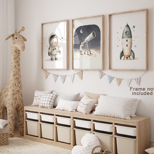 May include: Three framed illustrations of a child astronaut, a child looking through a telescope, and a rocket ship. The illustrations are in a light watercolor style with a white background. The frames are light wood and the illustrations are hung on a white wall. A woven giraffe figurine stands in the foreground.