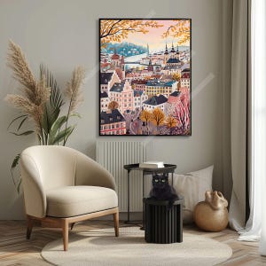 May include: A colourful painting depicting a European city with buildings, trees, and a river. The painting is framed in black and hangs on a wall in a room with a chair, a table, and a cat.