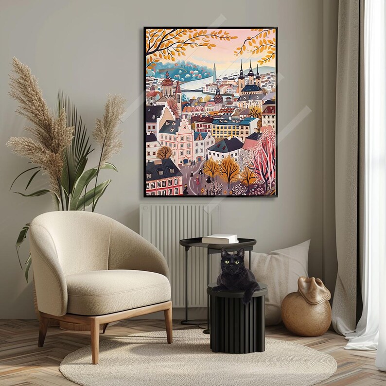 May include: A colourful painting depicting a European city with buildings, trees, and a river. The painting is framed in black and hangs on a wall in a room with a chair, a table, and a cat.