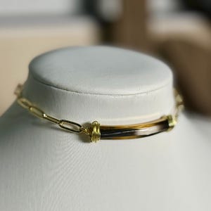 May include: A gold-toned chain necklace with a rectangular pendant featuring brown, black, and cream-colored stripes. The pendant is wrapped with gold wire. The necklace is displayed on a white mannequin.