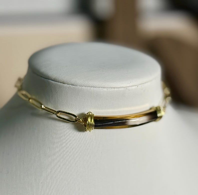 May include: A gold-toned chain necklace with a rectangular pendant featuring brown, black, and cream-colored stripes. The pendant is wrapped with gold wire. The necklace is displayed on a white mannequin.