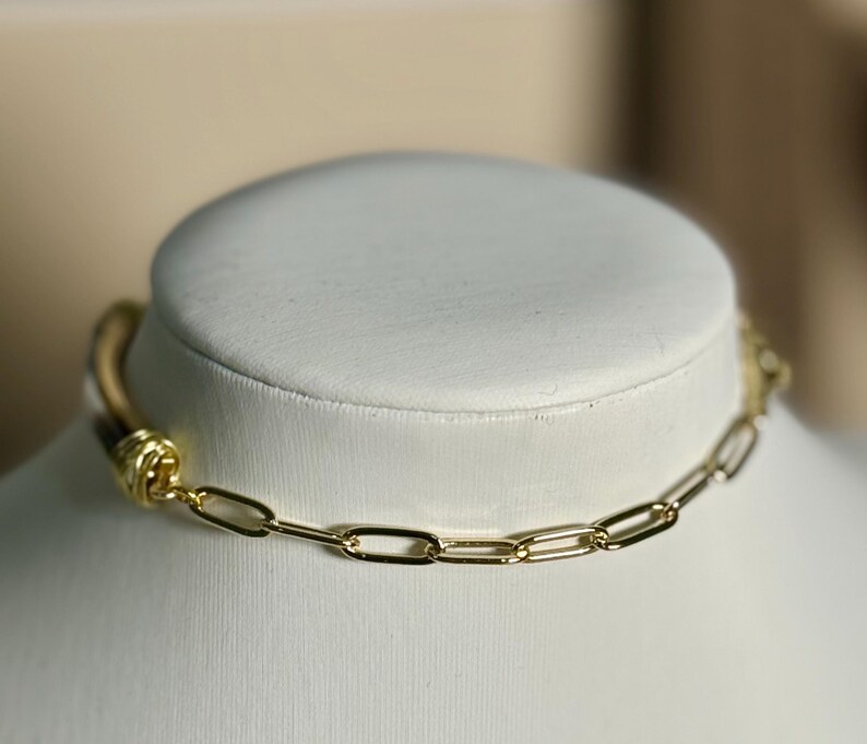 May include: A gold-toned chain necklace with a leather cord and a decorative knot. The necklace is displayed on a white bust, showcasing its design and craftsmanship. The chain links are rectangular.
