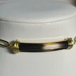 May include: A gold-toned chain necklace with a central, cylindrical pendant. The pendant is a mix of dark brown, tan, and white. The pendant is wrapped with gold wire at each end.