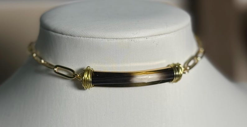 May include: A gold-toned chain necklace with a central, cylindrical pendant. The pendant is a mix of dark brown, tan, and white. The pendant is wrapped with gold wire at each end.