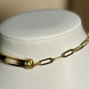 May include: A gold-toned chain necklace with a rectangular link design. The necklace features a central cylindrical element with brown and cream tones, wrapped in gold wire. The necklace is displayed on a white mannequin.