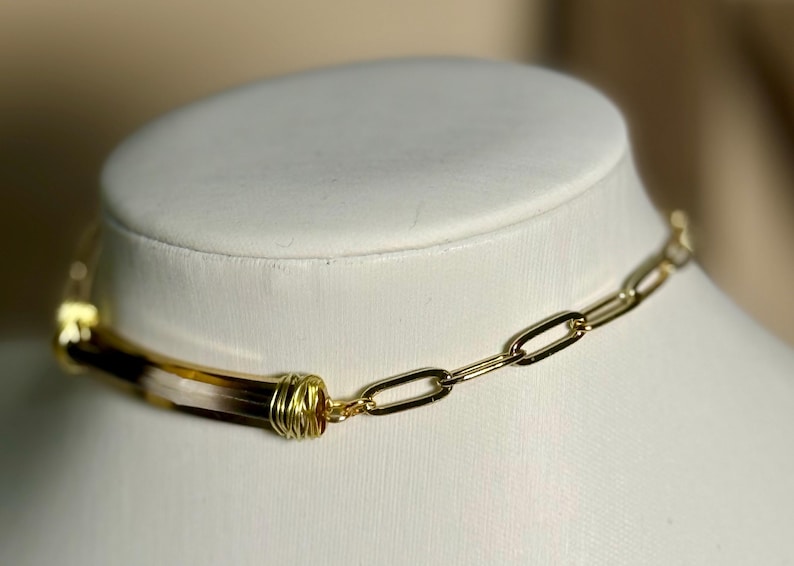 May include: A gold-toned chain necklace with a rectangular link design. The necklace features a central cylindrical element with brown and cream tones, wrapped in gold wire. The necklace is displayed on a white mannequin.