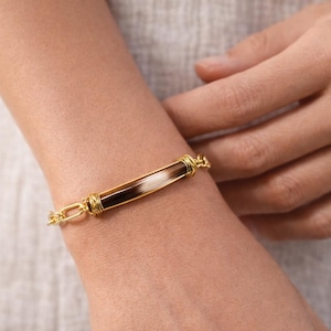 May include: A gold-toned bracelet with a rectangular centerpiece featuring a combination of white, brown, and black elements. The bracelet has a chain-link design and is worn on a wrist.