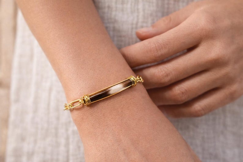 May include: A gold-toned bracelet with a rectangular centerpiece featuring a combination of white, brown, and black elements. The bracelet has a chain-link design and is worn on a wrist.