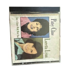 Puede incluir: Una caja de CD de música con una portada que muestra retratos de Patsy Cline y Loretta Lynn. El texto dice Patsy Cline, Loretta Lynn y Just A Closer Walk With Thee. El fondo es de color crema claro con un patrón de hojas sutil y repetitivo.