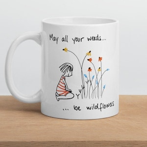 May include: White ceramic mug with a black line drawing of a girl and flowers. The text reads "May all your weeds... be wildflowers."