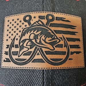 May include: A brown leather patch with a black silhouette of a fish and two fishing hooks. The fish is positioned over an American flag design with red and white stripes and white stars.