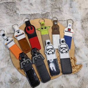 May include: A collection of keychains featuring Star Wars characters and symbols. The keychains have various designs, including a Stormtrooper helmet, Darth Vader, and Yoda. They are made of leather and have a metal keyring.